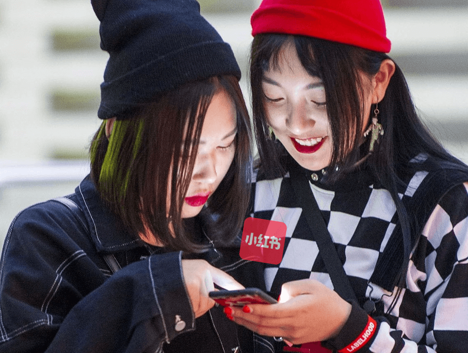 Two young women looking at a smartphone. One wears a black beanie and jacket; the other has a red beanie and checked shirt.